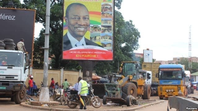 People pass in front of an electoral campaign poster for incumbent President Alpha Conde in Conakry, Guinea, Sept. 10, 2015.